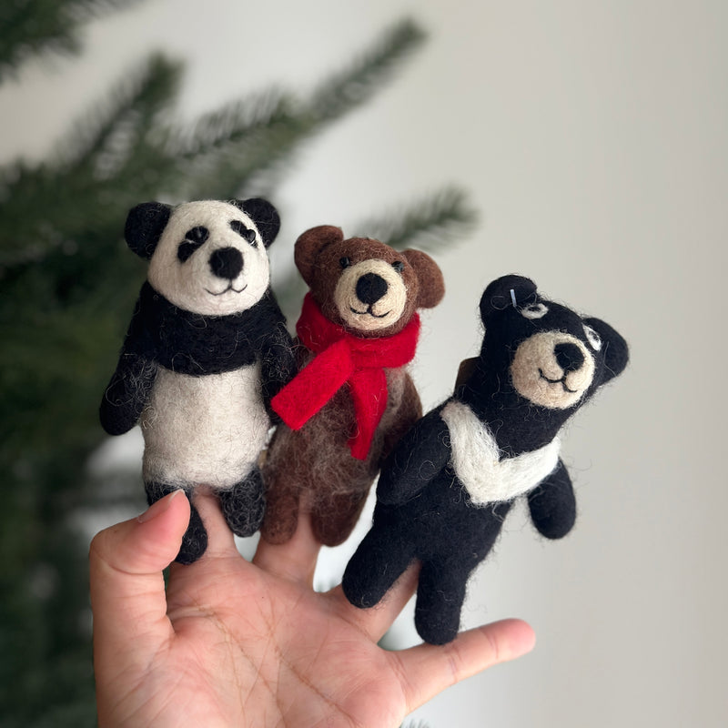 A handmade felt Kung Fu Panda finger puppet is playfully showcased on a female hand against a soft background with a partial view of a Christmas tree. This black and white felted finger puppet features distinct black ears and eye patches, along with a cheerful, smiling expression. Its textured wool felt material highlights the unique craftsmanship of this felt toy for kids.