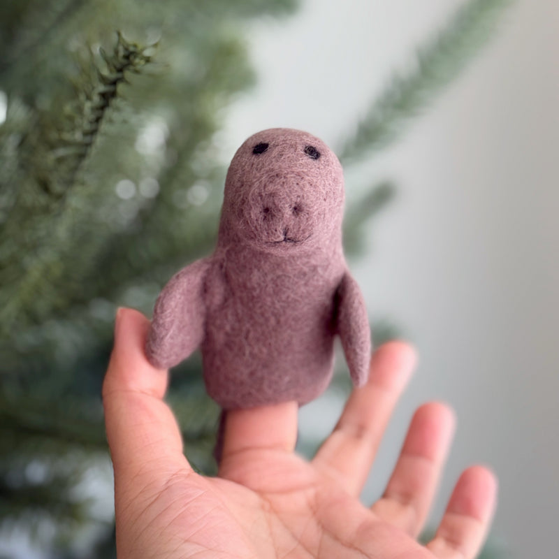 A handcrafted felt manatee finger puppet is gently held by a hand against a blurred background with a subtle Christmas tree. This unique felt toy for kids is a muted mauve color, featuring a rounded body, small flippers, and a simple face with black eyes, a textured snout, and a small mouth. Its design highlights its artisanal charm.