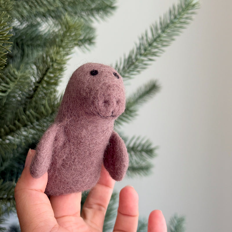 A handmade muted-purple felt manatee finger puppet rests against a blurred Christmas tree background. This charming felted finger puppet features a rounded body, tiny felt flippers, and a simple, expressive face with small, dark eyes, ideal for imaginative play.