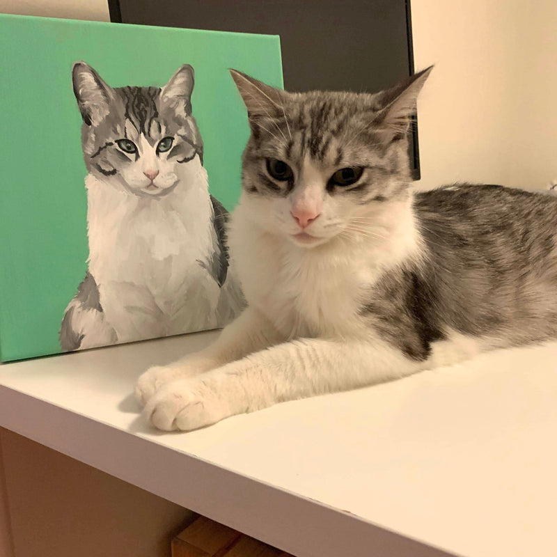 A gray and white felt cat finger puppet, named "Mochi," rests on a white surface, appearing soft and lifelike. Behind it, a painting of the same cat is displayed against a green backdrop, enhancing the domestic setting. This handcrafted felt finger puppet beautifully captures the cat's features, evoking warmth and connection.