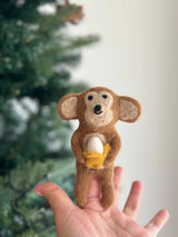 A handcrafted wool felt monkey finger puppet is held gently in a female hand. The light brown monkey features a warm smiling face, detailed with a small, peeled white and yellow banana clutched in its paws. This unique, ethically made felt puppet highlights skilled craftsmanship.
