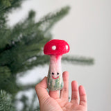A female hand holds a handmade wool felt mushroom finger puppet against a blurred Christmas tree backdrop. This whimsical mushroom puppet has a textured, bright red cap with white spots, and a stem that transitions from light pink to a greenish base, adorned with a small, cheerful smile.