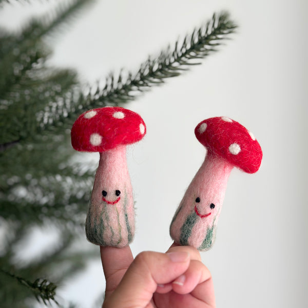 A handcrafted wool felt mushroom finger puppet is held gently in a female hand, set against a blurred Christmas pine tree. This unique felt mushroom finger puppet features a bright red cap with white spots, a light pink stem with subtle green stripes, and a small, cheerful smile, highlighting its whimsical design.