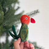 A handmade felt parrot finger puppet is held by a female hand against a soft white background, with a partially visible Christmas tree. This colorful felt parrot features an orange head, a white and red beak, a green body with yellow markings, and two green tail feathers.