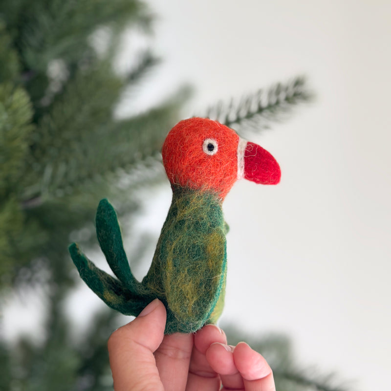 A handmade felt parrot finger puppet is held by a female hand against a soft white background, with a partially visible Christmas tree. This colorful felt parrot features an orange head, a white and red beak, a green body with yellow markings, and two green tail feathers.