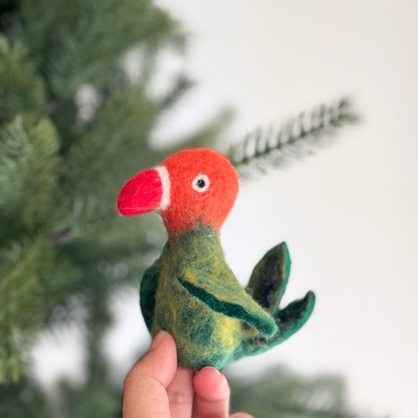 A playful felt parrot finger puppet is held by a female hand, showcasing its vibrant details. This cheerful handmade parrot features a red beak, an orange head, and a green body with small wings. A blurred Christmas tree in the background enhances the warm, festive mood.