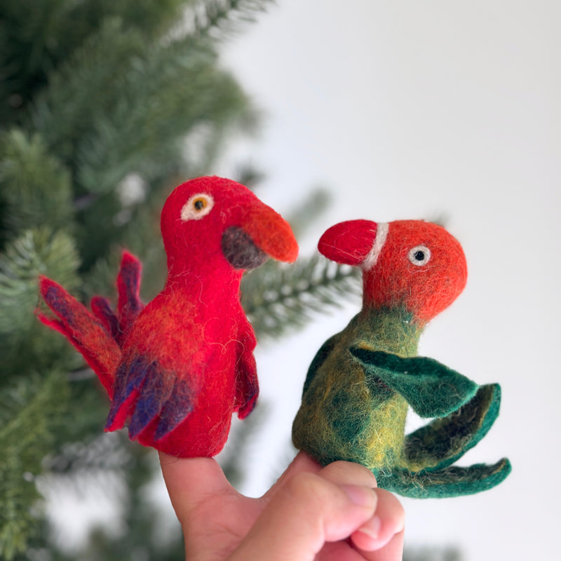 A handcrafted felt parrot finger puppet is playfully displayed in a close-up on a female hand, set against a blurred green Christmas tree backdrop. This vibrant felt parrot finger puppet features a predominantly red body accented with hints of orange and purple on its tail feathers and a notable beak. Its soft felt texture and unique design highlight its artisan craftsmanship, perfect for imaginative play.