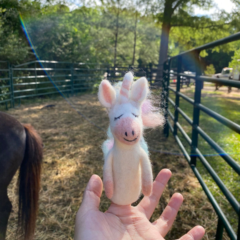 A handcrafted pastel unicorn felt finger puppet is held gently in a female hand, outdoors in a sunlit, grassy pasture. Behind the puppet, a metal fence and trees are visible, with a dark horse partially seen to the left. This whimsical needle-felted unicorn puppet is predominantly white, featuring pastel rainbow hair, a pink snout, and a charming winking eye, inviting imaginative play.