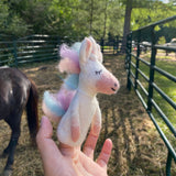 A female hand holds a pastel unicorn felt finger puppet outdoors in front of a green metal fence, with a horse visible in the background. This eco-friendly, handcrafted unicorn felted finger puppet is white with pastel rainbow hair in pink, blue, and white strands. It features a stitched closed eye and smiling mouth, showcasing its soft wool felt texture.