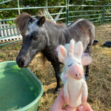 A handcrafted needle felted pastel unicorn finger puppet is held playfully in a female hand outdoors, with a real pony grazing in the background. This charming wool felt unicorn puppet is predominantly white with a pink muzzle, a winking eye, and a pastel rainbow mane, highlighting its whimsical, eco-friendly design.