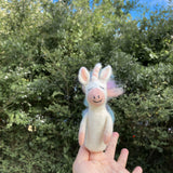 A handcrafted pastel unicorn felt finger puppet is held delicately in a female hand, set against blurred green foliage and a bright blue sky. This charming, eco-friendly wool felt unicorn features a white body, pastel rainbow hair, a pink snout, inner ears, and a pastel striped horn, contributing to its whimsical design with a winking eye.