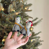 A handmade felt penguin finger puppet with headphones is presented by a female hand against a blurred Christmas tree background adorned with string lights, evoking a festive and playful mood. This unique felt penguin features a grey body, black feet, and a white face, accented by black headphones.