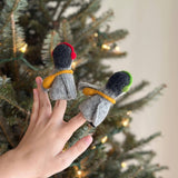 A unique felt penguin finger puppet with headphones is held on a female hand against a blurred background of a lit Christmas tree. This playful handmade felt penguin finger puppet features a gray body, a black head, and is accessorized with a yellow band representing a scarf and black headphones.