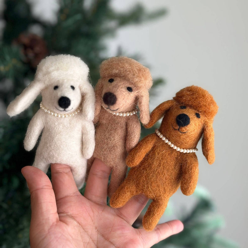 Multiple handcrafted felt poodle dog finger puppets are gently held in a female hand against a blurred, festive background. These charming puppets, available in white, beige, and brown, each feature a delicate plastic pearl necklace and fluffy hair, capturing the distinct charm of a poodle. Their soft texture highlights their sophisticated and fun design, emphasizing their craftsmanship.