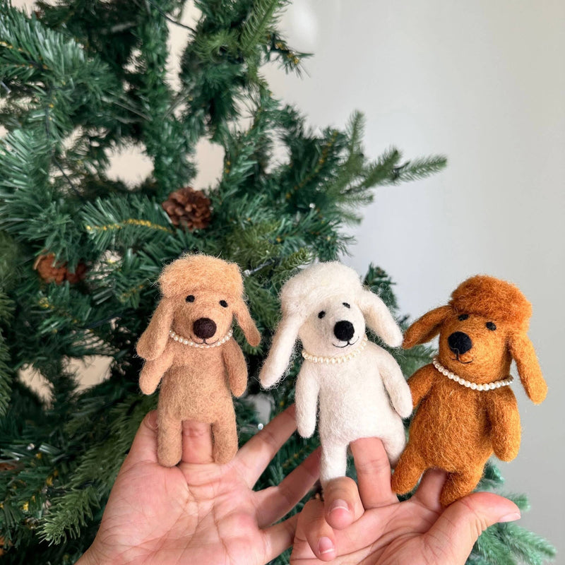 Three handcrafted felt poodle dog finger puppets are presented by a female hand against a festive Christmas tree adorned with pine cones. Each unique felt poodle dog puppet, in white, beige, and brown, features fluffy hair and a delicate plastic pearl necklace, perfect for imaginative finger play.