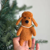 A handmade brown felt poodle finger puppet is held gently by a hand, presented against a soft, festive backdrop with a blurred Christmas tree. This charming felt poodle features fluffy brown hair and a delicate plastic pearl necklace, enhanced by warm, indirect lighting.