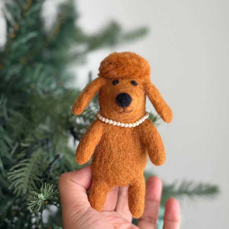 A handmade brown felt poodle finger puppet is held gently by a hand, presented against a soft, festive backdrop with a blurred Christmas tree. This charming felt poodle features fluffy brown hair and a delicate plastic pearl necklace, enhanced by warm, indirect lighting.