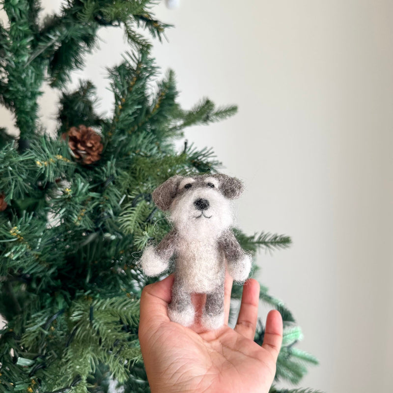 A gray and white Schnauzer felt finger puppet is held by a female hand, appearing in front of a festive Christmas tree. This handcrafted wool felt Schnauzer finger puppet features a textured design that mimics the dog's signature beard and bushy eyebrows. The cozy backdrop of a green Christmas tree, decorated with pine cones and lights, enhances the puppet's playful charm.