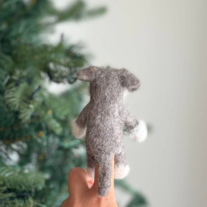 A wool felt Schnauzer finger puppet is held by a female hand, set against a cheerful backdrop with a blurred Christmas tree. This handcrafted puppet boasts a textured gray body, a tail, white paws, and the Schnauzer's signature beard and bushy eyebrows.