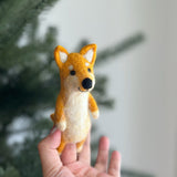 A handcrafted Shiba Inu wool felt finger puppet is gently held by a female hand, set against a blurred background suggesting a festive Christmas tree. The adorable Shiba Inu puppet has a light orange head with white markings, tiny black eyes, a black nose, and perky ears, showcasing intricate needle-felted details. Its body is cream, with white paws, completing its charming fox-like expression.