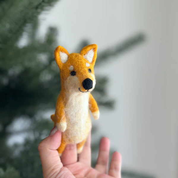 A handcrafted Shiba Inu wool felt finger puppet is gently held by a female hand, set against a blurred background suggesting a festive Christmas tree. The adorable Shiba Inu puppet has a light orange head with white markings, tiny black eyes, a black nose, and perky ears, showcasing intricate needle-felted details. Its body is cream, with white paws, completing its charming fox-like expression.