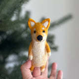 A handcrafted Shiba Inu felt finger puppet is gently held by a female hand, set against a softly blurred background of a Christmas tree and a plain wall. This unique wool felt finger puppet features the Shiba Inu’s distinctive fox-like expression with intricate needle-felted details. It is predominantly orange with white accents on its chest, paws, and inner ears, detailed with black eyes and nose.