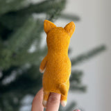 A handmade wool felt Shiba Inu finger puppet is held by a female hand against a blurred Christmas tree background. This vibrant yellow dog finger puppet features a textured surface, upright ears, and white paws, highlighting its charming, handcrafted details.