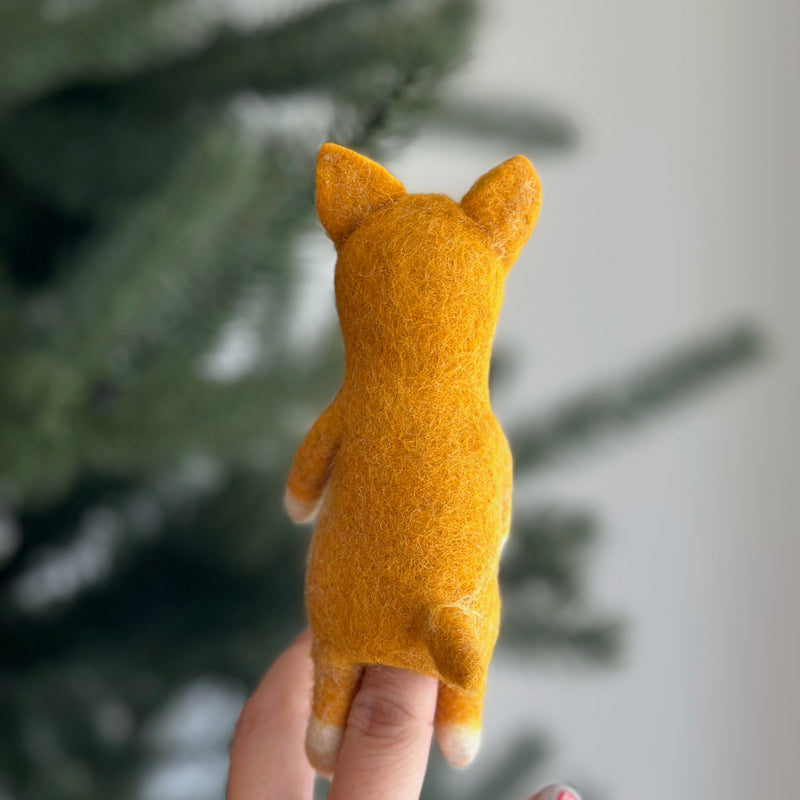 A handmade wool felt Shiba Inu finger puppet is held by a female hand against a blurred Christmas tree background. This vibrant yellow dog finger puppet features a textured surface, upright ears, and white paws, highlighting its charming, handcrafted details.