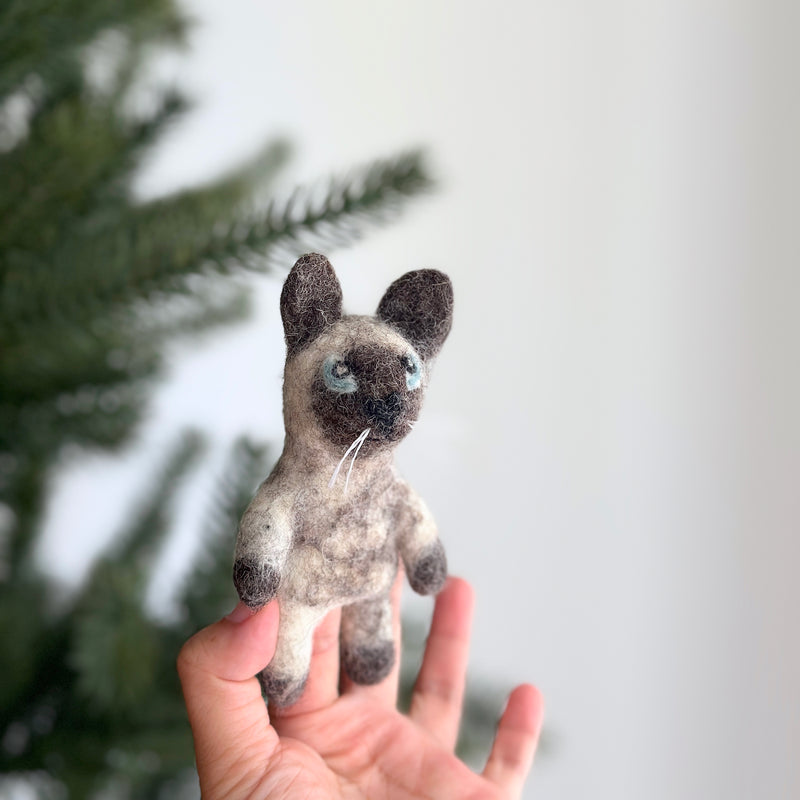 A handcrafted felt Siamese cat finger puppet is held by a female hand against a bright, cheerful backdrop with a blurred Christmas tree. The plush felt puppet features a light tan body with dark brown markings on its face, ears, and paws, blue eyes, and white whiskers. This unique, fair-trade felt toy for kids is ethically made, adding charm to playtime.