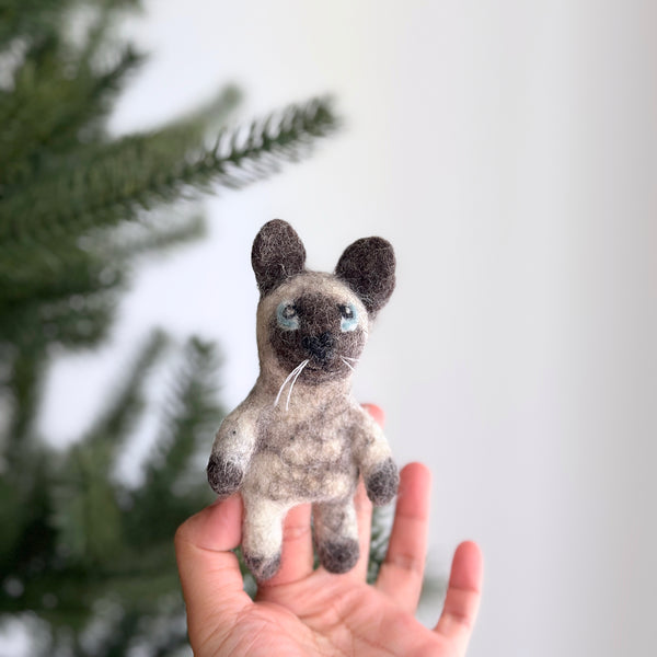 A handmade Siamese cat felt finger puppet is held in a female hand, with a blurred Christmas tree in the background, creating a festive mood. This unique, needle felted puppet showcases the distinctive pointed coloration of a Siamese cat, featuring darker ears, paws, and face contrasting its lighter body. It has charming blue eyes and delicate white whiskers. Crafted from plush felt by a Fair Trade artisan collective in Nepal, this eco-friendly toy supports imaginative play.