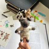 A handmade felt finger puppet shaped like a Siamese cat, resting on a person's hand, against a warm and inviting background of books and a small stuffed animal. The puppet features soft texture and distinctive markings typical of a Siamese cat, made from wool felt. A delightful toy for kids, this unique finger puppet is perfect for imaginative play.