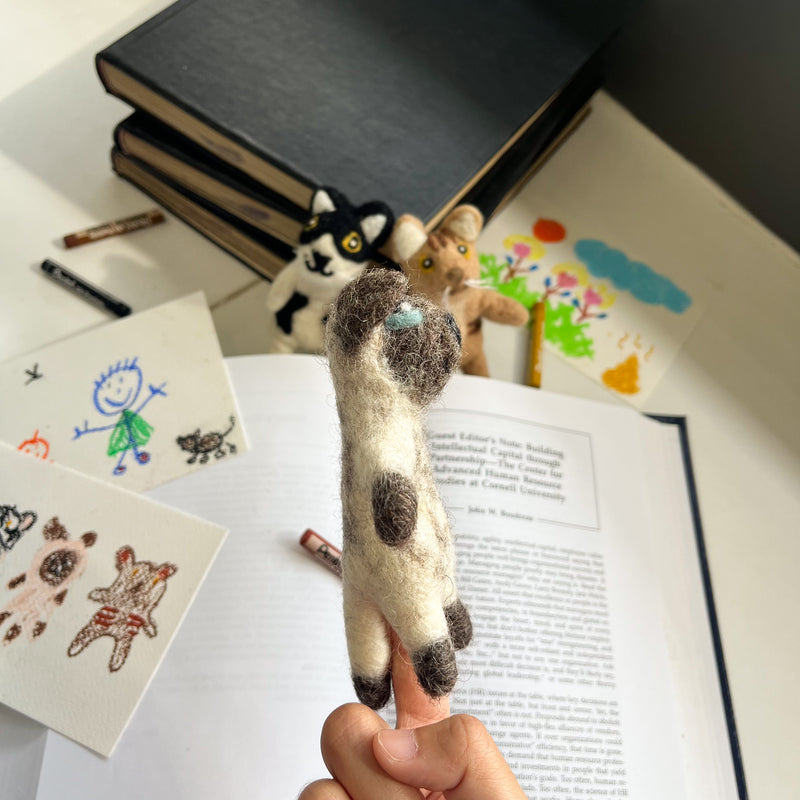 A plush felt siamese cat finger puppet is presented, worn on a finger against a backdrop of children's drawings and open books. This handcrafted felt kids toy displays a light brown body with dark brown markings on its paws, face, and tail. Lovingly made by a Fair Trade artisan collective in Nepal, it embodies ethical and eco-friendly craftsmanship for creative play.