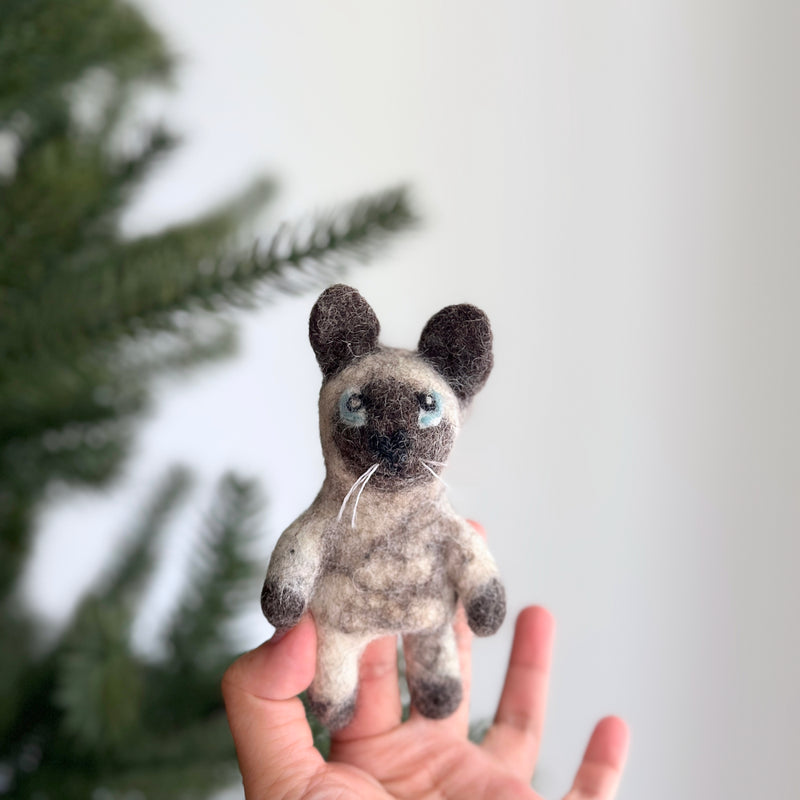 A handcrafted felt Siamese cat finger puppet is held closely in a female hand, playfully set against a blurred Christmas tree background. This charming felt puppet features a grey face with a white section extending from its nose to its chest, complemented by dark grey ears, paws, and tail. Blue eyes and white whiskers add to its adorable feline look. This unique, handcrafted toy is made with ethical and eco-friendly practices.