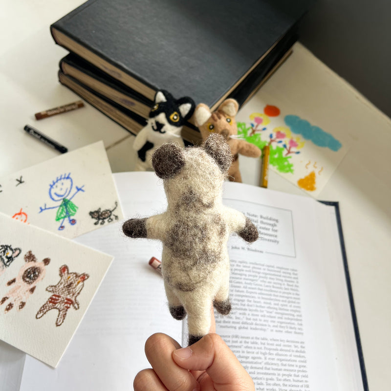A handmade wool felt Siamese Cat finger puppet is held by a hand, presented in a playful, educational setting with an open book, drawings, and crayons. Two other cat felt finger puppets are softly blurred in the background. This uniquely crafted felt toy, made from soft, textured wool felt by a Fair Trade artisan collective in Nepal, fosters creative play.