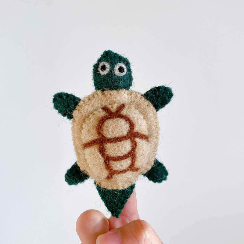 A wool felt turtle finger puppet is playfully displayed on a person's finger against a bright white background. This charming, needle-felted turtle is handmade of 100% wool, featuring a green head, four green legs, and a green tail. Its 3D shell is distinctively beige with a brown design.