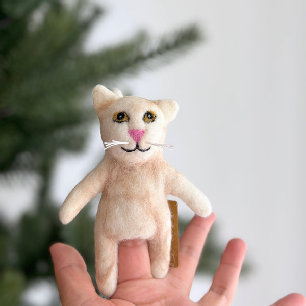 A handcrafted Tuxedo Cat felt finger puppet is gently held in a female hand against a blurred, festive backdrop featuring a green Christmas tree branch. This unique felt finger puppet showcases soft black and white felt, delicate whiskers, small ears, and a stitched smile. Crafted from plush, eco-friendly felt by a fair-trade artisan collective in Nepal, it offers a soft, tactile, and engaging play experience.