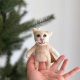 A handmade felt Tuxedo Cat finger puppet is presented gently in a human hand, with a softly blurred green Christmas tree in the background. This meticulously crafted felt cat features small upright ears, black eyes, a pink nose, and delicate white whiskers, all made from plush felt. This unique puppet is a product of ethical and eco-friendly practices by a Fair Trade artisan collective in Nepal.