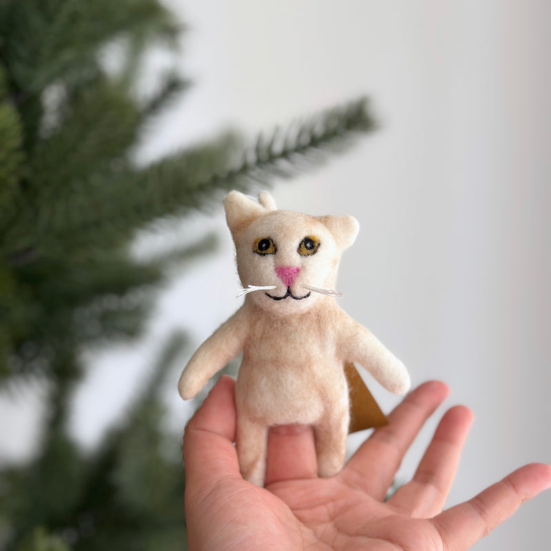 A handmade felt Tuxedo Cat finger puppet is presented gently in a human hand, with a softly blurred green Christmas tree in the background. This meticulously crafted felt cat features small upright ears, black eyes, a pink nose, and delicate white whiskers, all made from plush felt. This unique puppet is a product of ethical and eco-friendly practices by a Fair Trade artisan collective in Nepal.