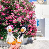A female hand holds two charming felt duck finger puppets, dressed as a couple on honeymoon, against a vibrant floral and traditional architectural background. One handmade felt duck wears a pink hat, while the other features a black hat and scarf, creating a delightful, travel-inspired scene.