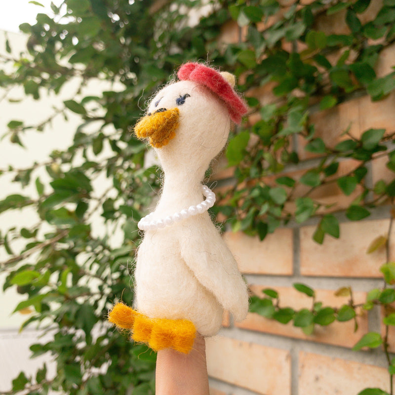 A handmade wool felt duck finger puppet, from the 'Honeymoon' collection, is worn on a female hand against green foliage and a brick wall. This off-white felt duck has a textured finish, bright yellow beak and feet. Adorned with a red felt beret and a string of white pearls, this charming handcrafted felt kids toy reflects its delightful honeymoon attire.