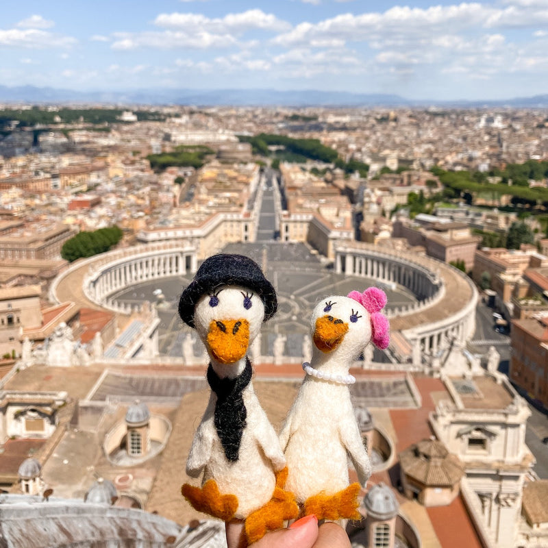 A pair of handcrafted felt duck couple finger puppets are held up against a sunny, sprawling European cityscape. The male felt duck finger puppet wears a black hat and tie, while the female felt duck finger puppet sports a pink hair accessory and pearl necklace. Both charming white duck puppets feature orange beaks and feet, embodying a delightful honeymoon theme.
