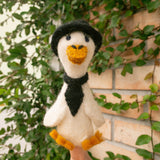 A handcrafted wool felt duck finger puppet from the 'Duck Couple on Honeymoon' set is held by a female hand against a vibrant brick wall adorned with green leaves. This charming off-white felt duck puppet wears a black hat and tie, complemented by bright yellow accents on its beak and feet. The soft, handmade felt toy is perfect for imaginative play and storytelling.