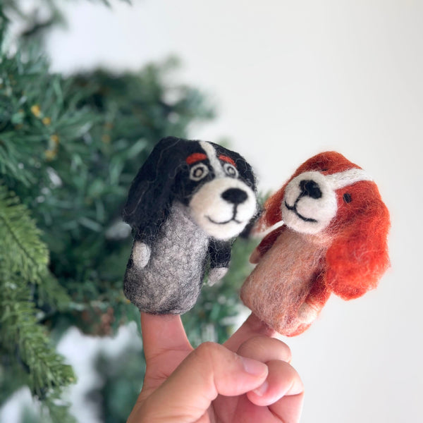 Two handcrafted Cavalier King Charles Spaniel felt finger puppets are held in a hand against a blurred Christmas tree background. One puppet depicts a black Cavalier King Charles Spaniel with black ears, a grey body, and white markings. The second is an orange Cavalier King Charles Spaniel with orange ears, an orange body, and white markings, both featuring intricate felt detailing for imaginative play.