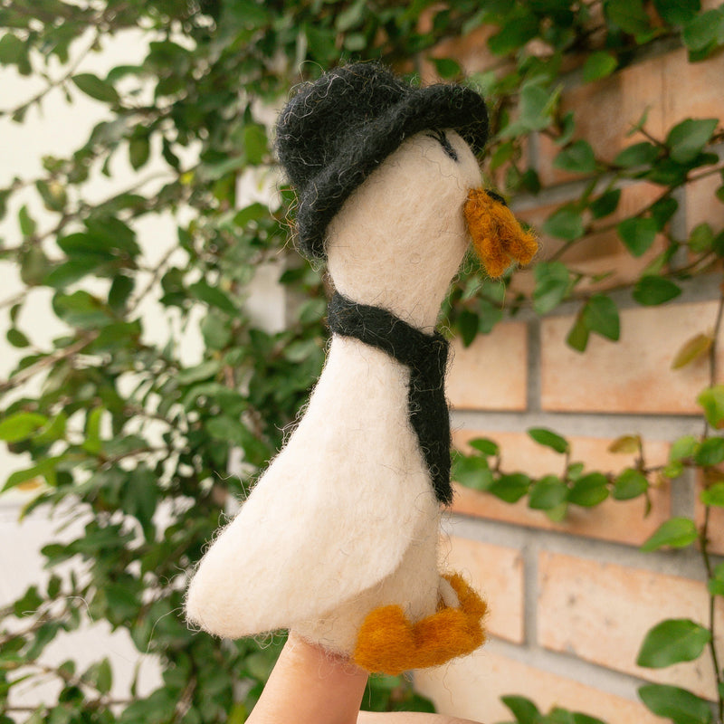 A handcrafted felt duck finger puppet, expertly made, is presented on a finger against green vines and a brick wall. This charming white duck has a fluffy texture, orange beak and feet, and is adorned with a black hat and scarf. This whimsical handmade felt duck finger puppet showcases detailed craftsmanship, inspiring creative imaginative play while supporting artisans.