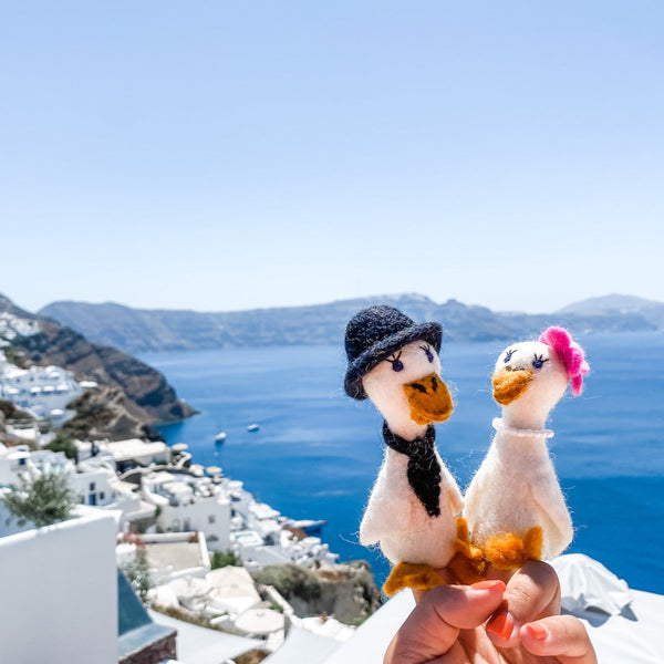 Two handcrafted felt duck finger puppets are held by a female hand against a bright coastal background of blue sky, sea, and whitewashed buildings. The charming felt duck couple, designed for a "honeymoon" theme, are white with orange beaks and feet. One duck wears a black top hat and tie, while the other features a pink flower and pearl necklace, showcasing their unique and intricate craftsmanship.