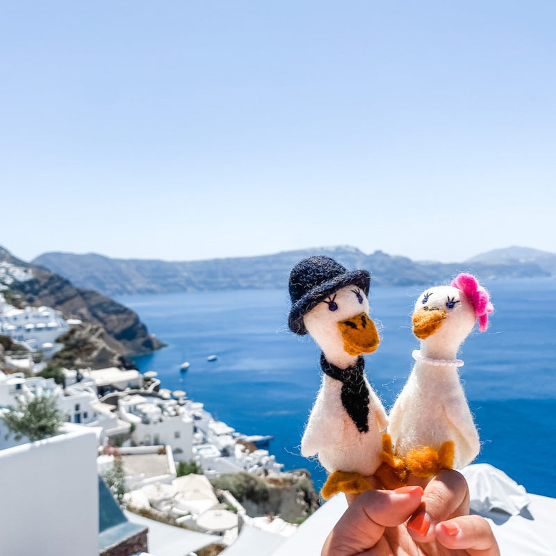 Two handcrafted felt duck finger puppets are held by a female hand against a bright coastal background of blue sky, sea, and whitewashed buildings. The charming felt duck couple, designed for a "honeymoon" theme, are white with orange beaks and feet. One duck wears a black top hat and tie, while the other features a pink flower and pearl necklace, showcasing their unique and intricate craftsmanship.