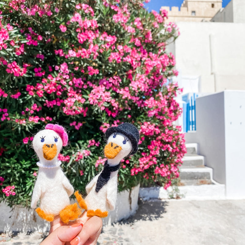 Two handcrafted felt duck finger puppets, portraying a whimsical duck couple, are held by a female hand against a vibrant backdrop of pink flowers and white buildings. The white felt duck on the left wears a pink hat and white collar, while the one on the right sports a black hat and scarf. Both adorable duck puppets feature orange beaks and feet, highlighting their charming, expertly crafted details.