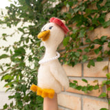 A handcrafted wool felt duck finger puppet is held by a female hand, set against lush green vines climbing a brick wall. This soft, off-white duck features a bright orange beak and feet. A red beret sits jauntily on its head, complemented by a delicate pearl necklace, reflecting the 'Honeymoon' theme. This exquisitely crafted puppet embodies whimsical design.