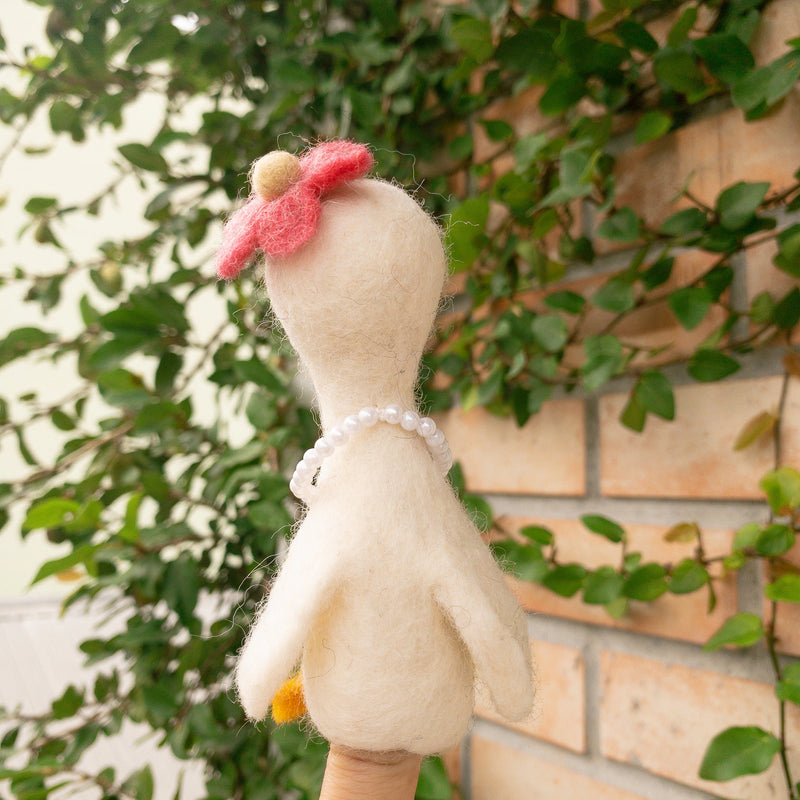 A handcrafted felt duck finger puppet, part of the 'Duck Couple on Honeymoon' set, is held by a female hand against a brick wall partially covered with green vines. This cream-colored wool felt duck puppet features a red flower atop its head and a strand of white beads around its neck. Its fuzzy texture adds to its whimsical charm.