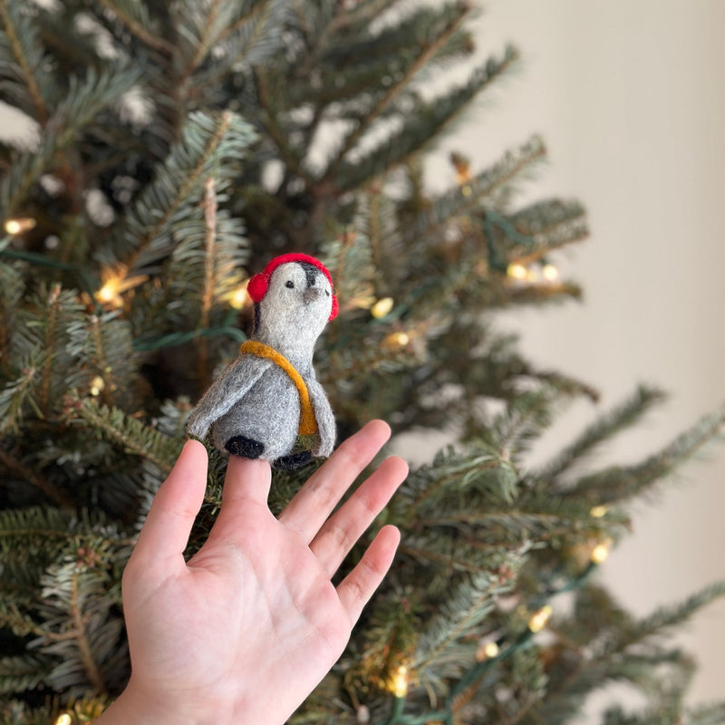 A handcrafted wool felt penguin finger puppet sits on a female finger, displayed against a blurred Christmas tree adorned with warm lights. This grey and white felt penguin finger puppet features playful red headphones and a yellow scarf, highlighting its unique design and supporting female artisans in Nepal.